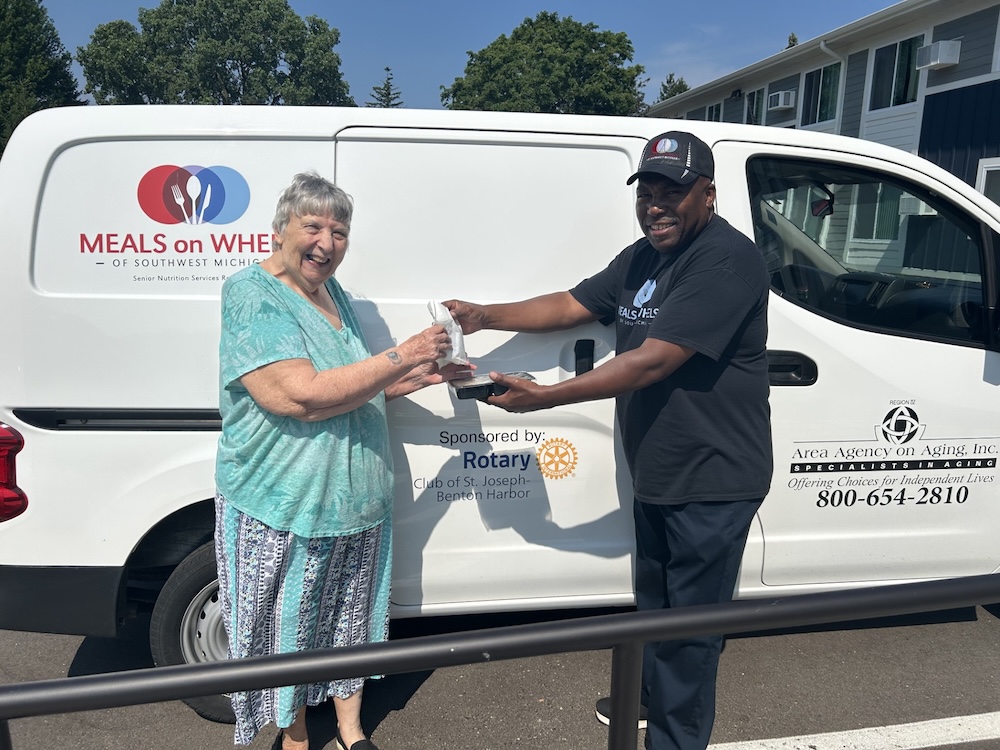 Smiling woman receives a meal from a man in front of a Meals on Wheels van in sunny weather.